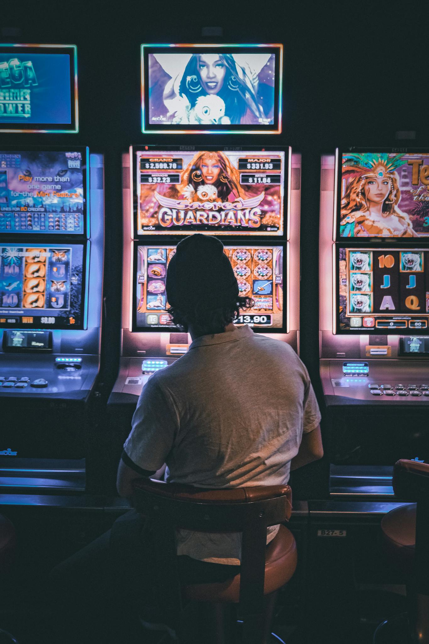Adult male seated in a dimly lit casino engaging with vibrant slot machines at night.